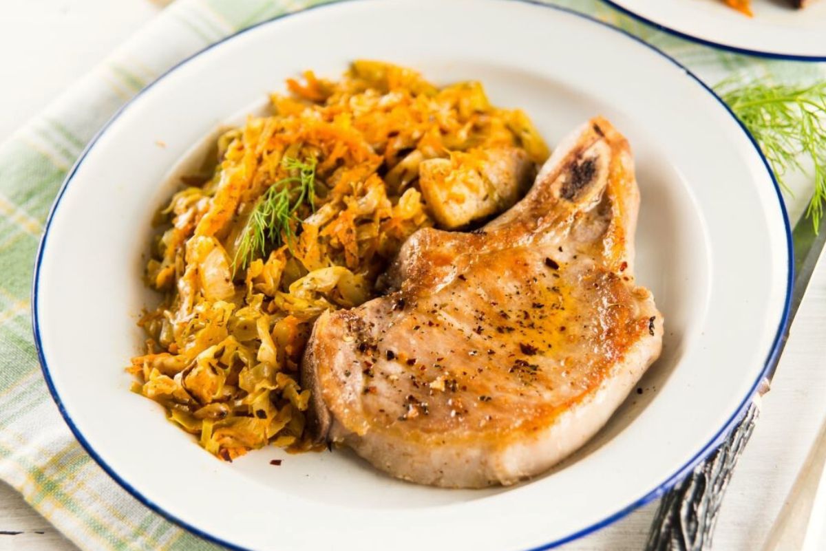High protein skillet pork chop with a golden seared crust served on a white plate with sautéed cabbage, shredded carrots, and tender vegetables. Juicy bone-in pork chop seasoned with black pepper, paired with cooked veggies for a simple one skillet dinner.