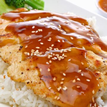 High protein teriyaki chicken breast glazed with shiny brown teriyaki sauce and sprinkled with sesame seeds, served over fluffy white rice on a white plate. Steamed broccoli sits in the background, with close-up food photography highlighting the sauce and chicken texture.