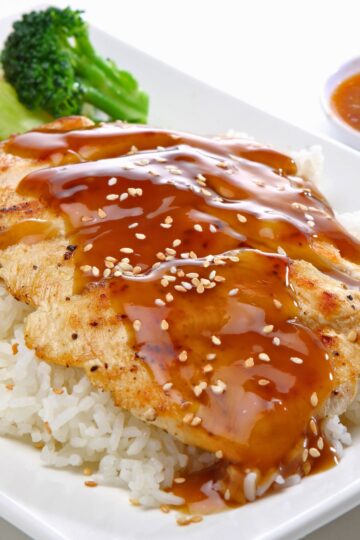 High protein teriyaki chicken breast glazed with shiny brown teriyaki sauce and sprinkled with sesame seeds, served over fluffy white rice on a white plate. Steamed broccoli sits in the background, with close-up food photography highlighting the sauce and chicken texture.