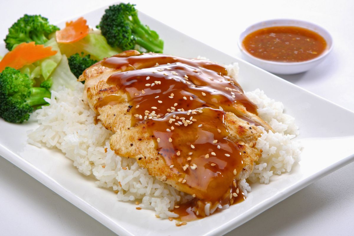 High protein teriyaki chicken breast sliced and drizzled with glossy brown teriyaki sauce, topped with sesame seeds, served over white rice on a square white plate. Steamed broccoli, carrots, and cabbage on the side, with a small bowl of extra teriyaki sauce in the background.