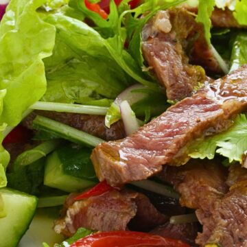 Close-up photograph of a Thai steak salad showing sliced medium-rare grilled beef with visible pink center arranged over fresh mixed greens including lettuce and arugula, sliced cucumbers, red bell pepper pieces, and sliced green onions. The beef appears glazed and glistening, and the salad ingredients are vibrant and fresh.