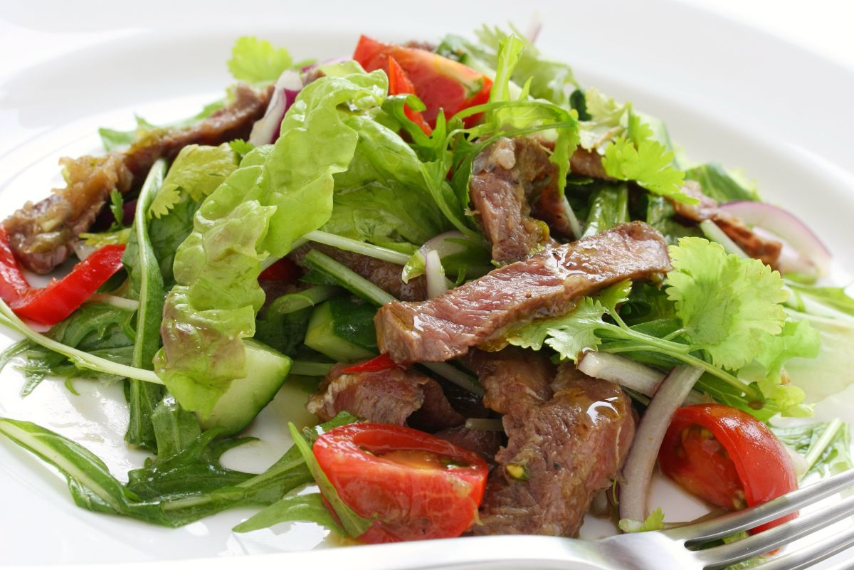 Thai steak salad served on a white plate, featuring sliced medium-rare grilled beef arranged over a bed of mixed greens including lettuce, arugula, and fresh cilantro, with sliced cucumbers, red bell peppers, red onions, green onions, and cherry tomato halves. A silver fork is visible at the edge of the plate on the right side.