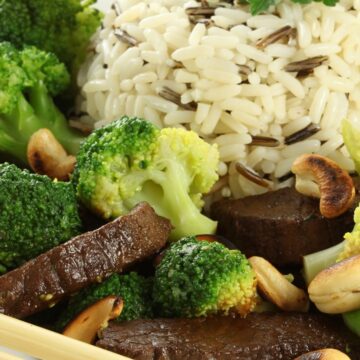 High protein Vietnamese beef bowl with sliced beef, steamed broccoli, toasted cashews, and a mound of white and wild rice on a white plate. Chopsticks rest along the edge, with fresh parsley on top of the rice.
