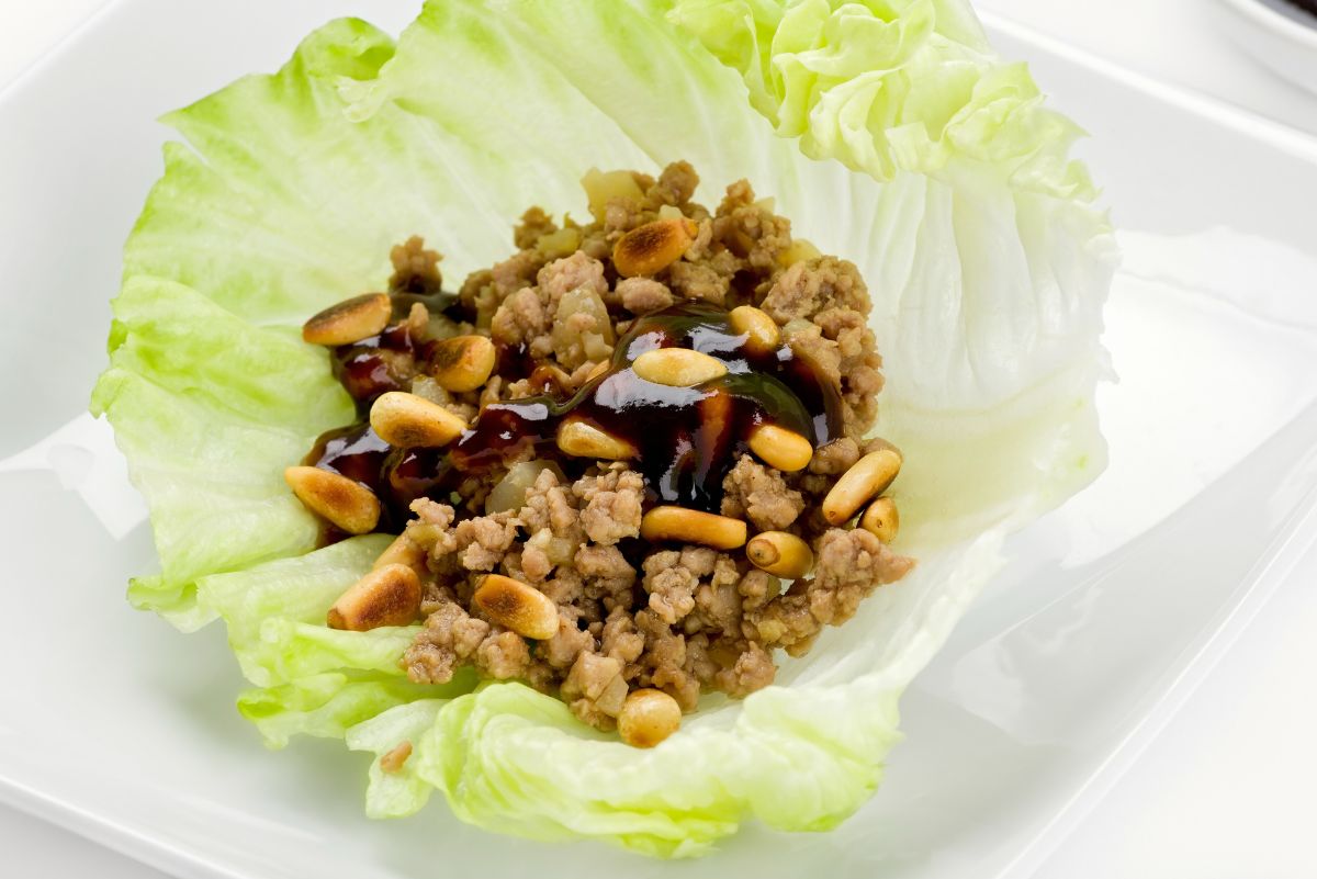 Hoisin ground beef lettuce wrap served on a white plate, filled with seasoned ground beef and diced onions, topped with toasted peanuts and drizzled with glossy hoisin sauce. The savory beef mixture is nestled in a crisp pale green lettuce leaf, shown from above.