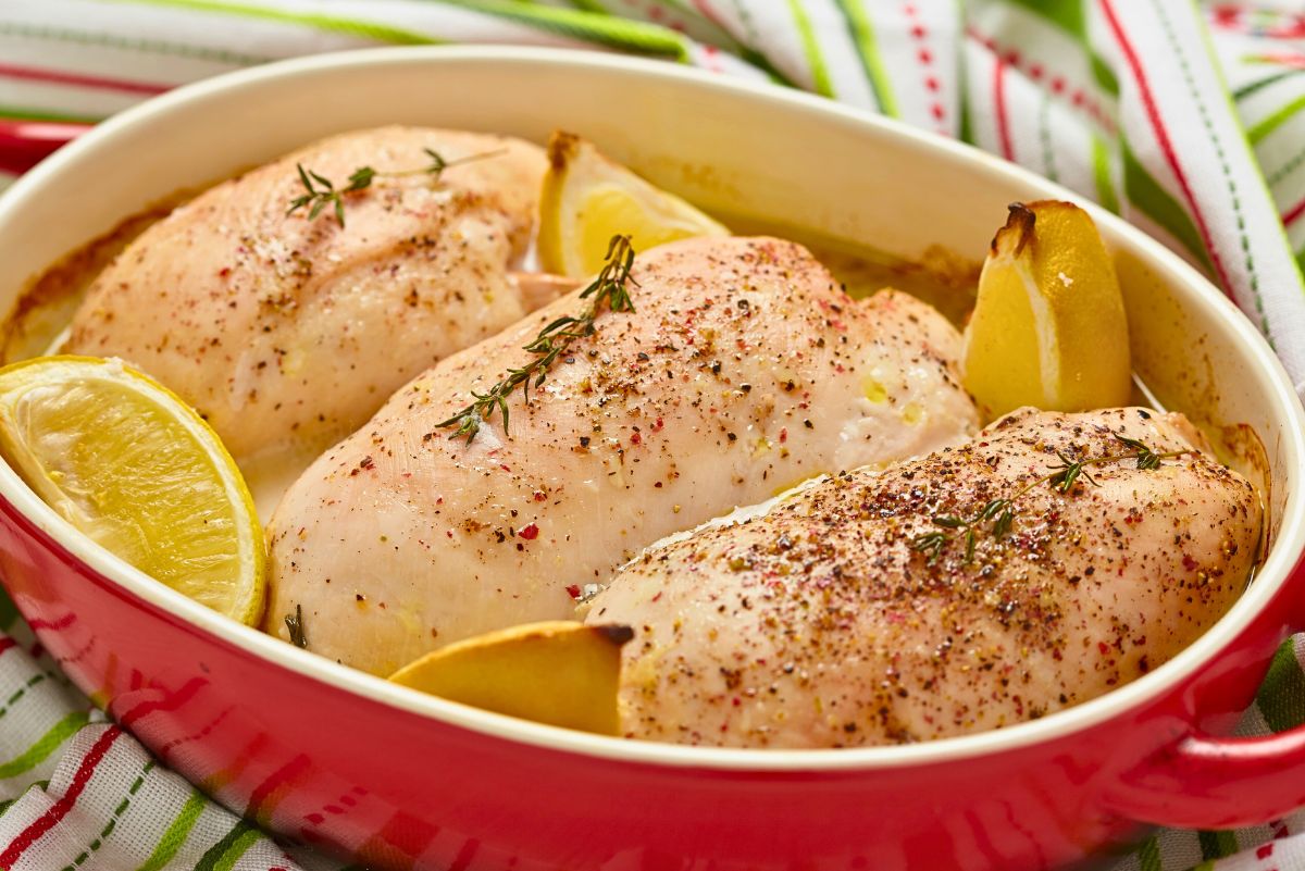 Red baking dish filled with lemon garlic baked chicken breasts, sprinkled with black pepper and herbs, with roasted lemon wedges tucked around the chicken on a striped kitchen towel background.