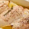 Close-up of seasoned lemon garlic baked chicken breasts in a red baking dish with lemon wedges and fresh herbs, ready to cook. Text on image: “nestinglane.com”.