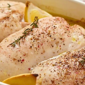 Close-up of seasoned lemon garlic baked chicken breasts in a red baking dish with lemon wedges and fresh herbs, ready to cook. Text on image: “nestinglane.com”.