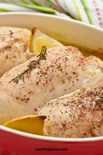 Close-up of seasoned lemon garlic baked chicken breasts in a red baking dish with lemon wedges and fresh herbs, ready to cook. Text on image: “nestinglane.com”.