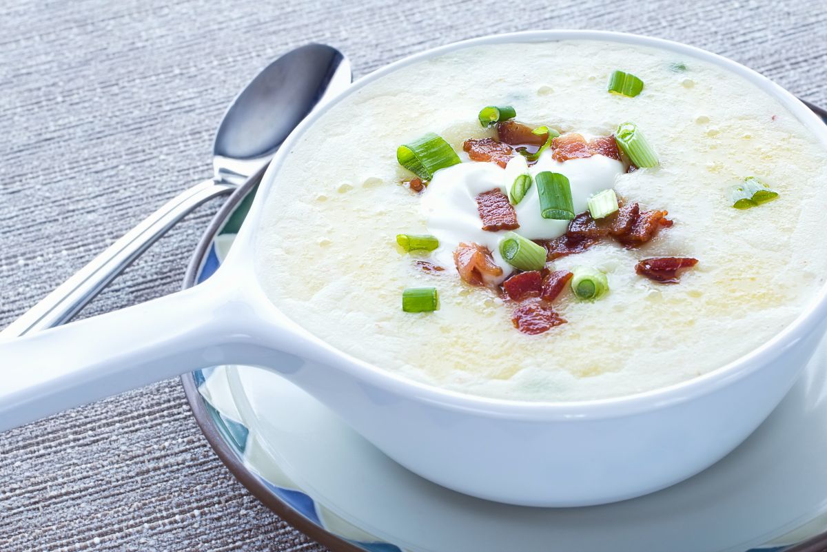 Creamy loaded potato soup in a white handled soup bowl on a saucer, topped with chopped turkey bacon, sliced green onions, and a small dollop of sour cream. Thick, smooth potato soup with visible bacon pieces and scallions, styled as a healthier comfort food dish with a spoon on a textured gray surface.