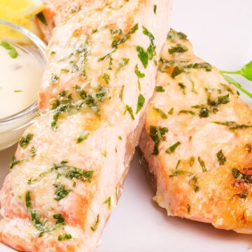 High protein parmesan herb salmon fillets baked until lightly golden and topped with melted cheese and chopped herbs, served on a white plate with a lemon wedge and a small bowl of creamy sauce on the side.