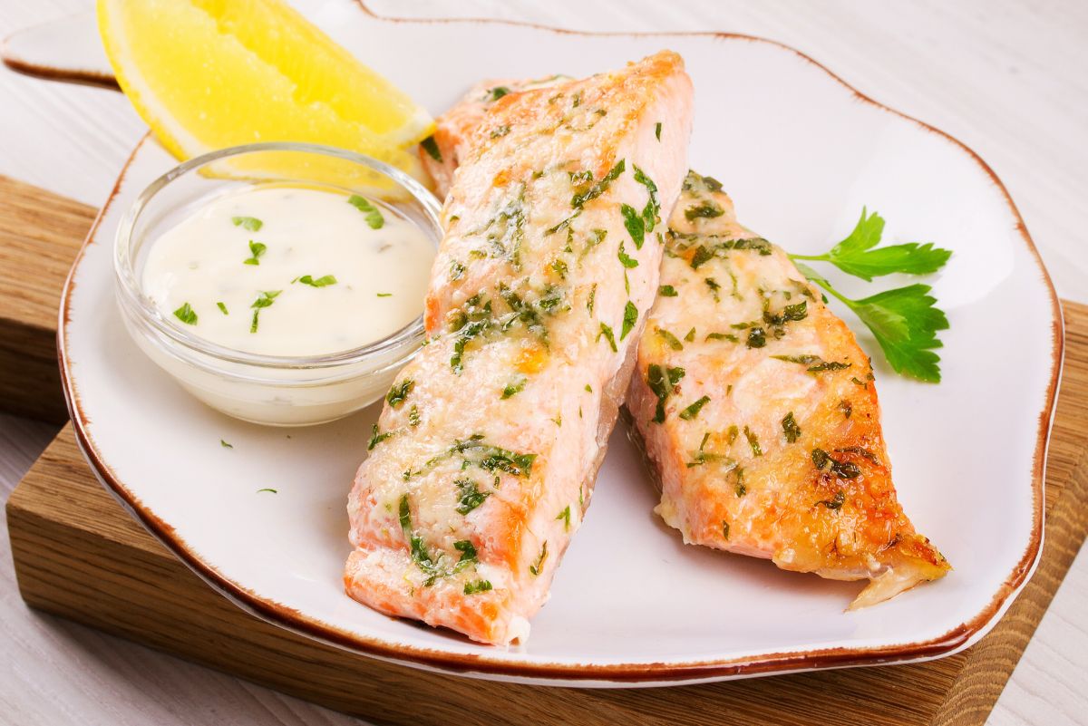 High protein parmesan herb salmon fillets baked until lightly golden, topped with melted cheese and chopped herbs. Served on a white plate with a lemon wedge, parsley garnish, and a small glass bowl of creamy sauce on a wooden board.