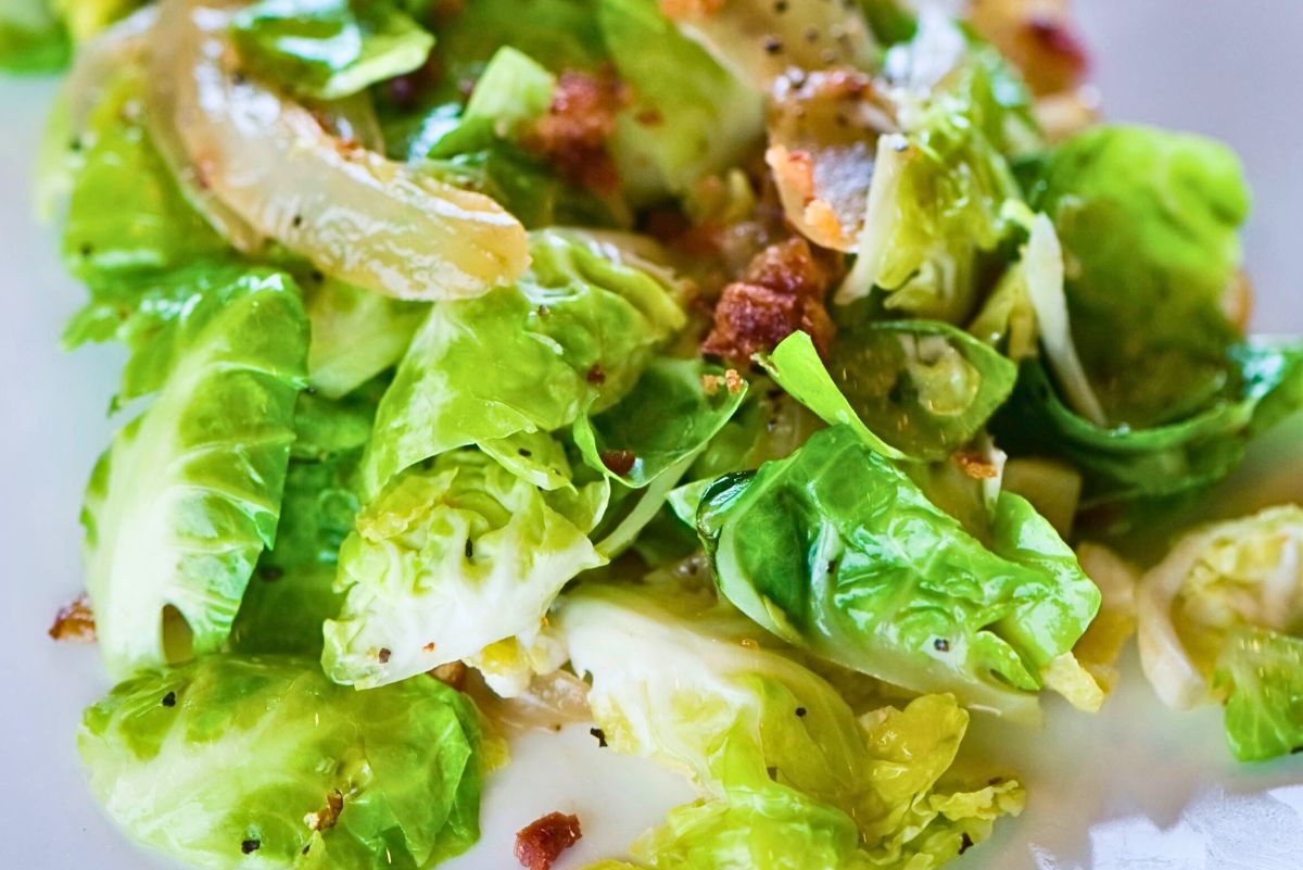 Close-up of a savory Brussels sprouts salad with tender bright green Brussels sprouts, crispy bacon bits, and sautéed onion slices coated in a glossy dressing. The sprouts look lightly caramelized with visible black pepper and bacon pieces on a white plate.