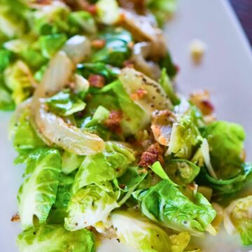 Savory Brussels sprouts salad with chopped bright green Brussels sprouts, crispy bacon bits, and sautéed onion slices tossed in a glossy dressing. Close-up view shows tender sprouts, bits of bacon, and cracked black pepper on a white plate, highlighting a healthy Brussels sprouts salad.