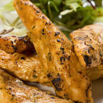 Close-up of smoky chipotle peanut chicken tenders with a golden-brown, lightly charred exterior and visible seasoning. Juicy grilled chicken strips rest on a white plate with mixed leafy greens in the background, highlighting spicy peanut chipotle chicken tenders with a savory, homemade finish.