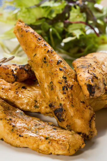 Close-up of smoky chipotle peanut chicken tenders with a golden-brown, lightly charred exterior and visible seasoning. Juicy grilled chicken strips rest on a white plate with mixed leafy greens in the background, highlighting spicy peanut chipotle chicken tenders with a savory, homemade finish.