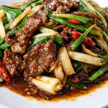 Close-up of beef and green onion stir fry on a white plate. Sliced beef, green onions, white onion, and red chili peppers are coated in a glossy brown sauce on a wooden table. High protein beef stir fry with green onions and chili.