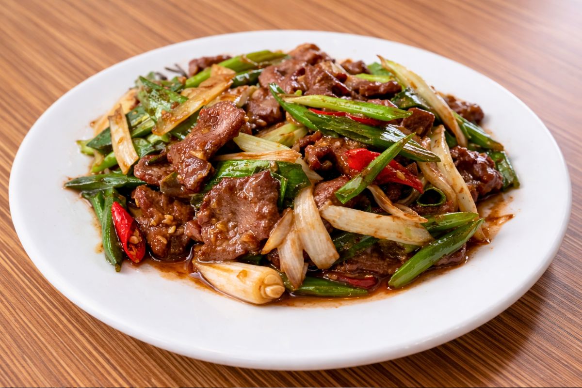 Beef and green onion stir fry served on a white plate. Tender sliced beef, green onions, white onion, and red chili peppers are coated in a glossy brown sauce on a wood table. High protein beef stir fry dinner.