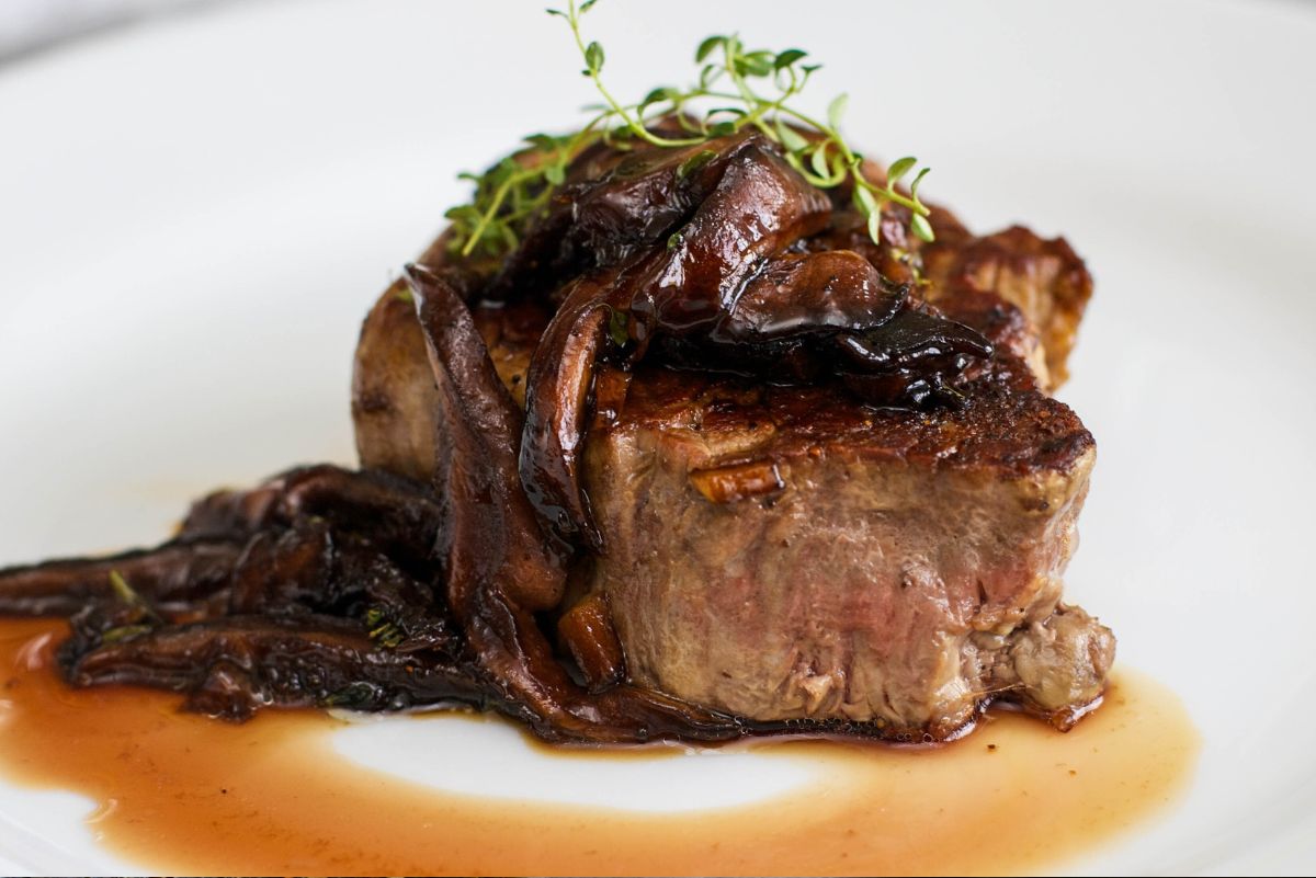 Filet mignon with mushroom wine sauce plated on a white dish. Thick seared steak topped with glossy sautéed mushrooms and thyme, with extra mushrooms and a light brown pan sauce around the steak. Elegant steak dinner close-up with rich, savory detail.