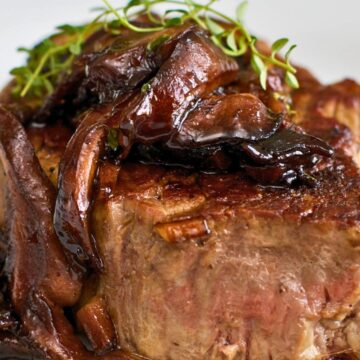 Filet mignon with mushroom wine sauce on a white plate, shown in a close-up food photo. Thick seared steak topped with glossy sautéed mushrooms and fresh thyme, with rich brown pan sauce pooling around the bottom. Gourmet steak dinner, restaurant-style plating.