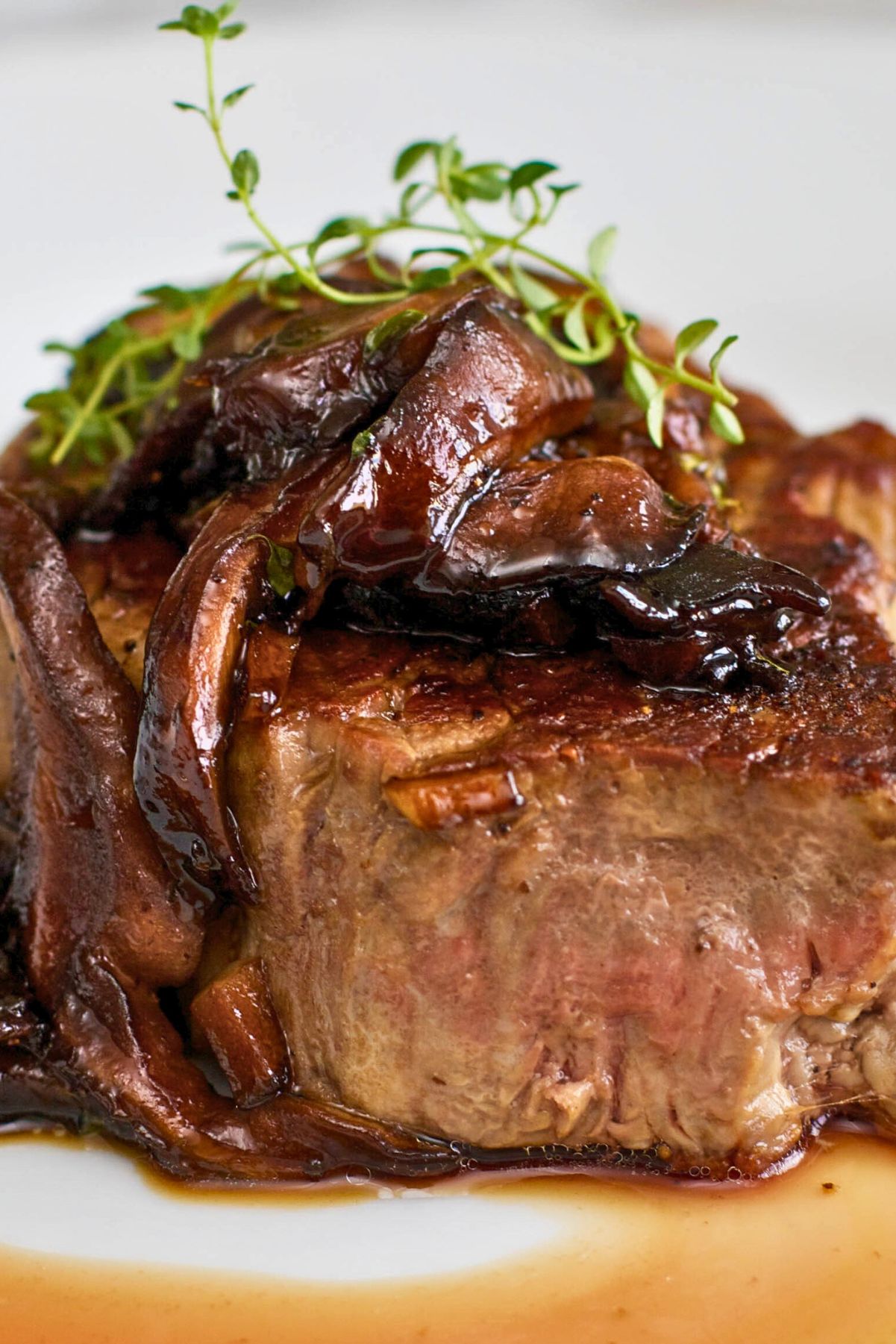 Filet mignon with mushroom wine sauce on a white plate, shown in a close-up food photo. Thick seared steak topped with glossy sautéed mushrooms and fresh thyme, with rich brown pan sauce pooling around the bottom. Gourmet steak dinner, restaurant-style plating.