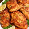 Crispy skillet Parmesan chicken on a white platter with lemon wedges and fresh parsley. Golden brown breaded chicken breasts are arranged close together in a plated food photo with a rustic wood background, showing a crunchy coating and herb garnish.