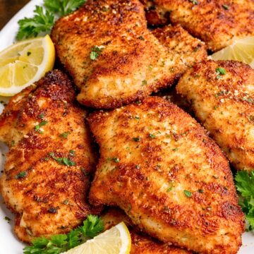Crispy skillet Parmesan chicken on a white platter with lemon wedges and fresh parsley. Golden brown breaded chicken breasts are arranged close together in a plated food photo with a rustic wood background, showing a crunchy coating and herb garnish.