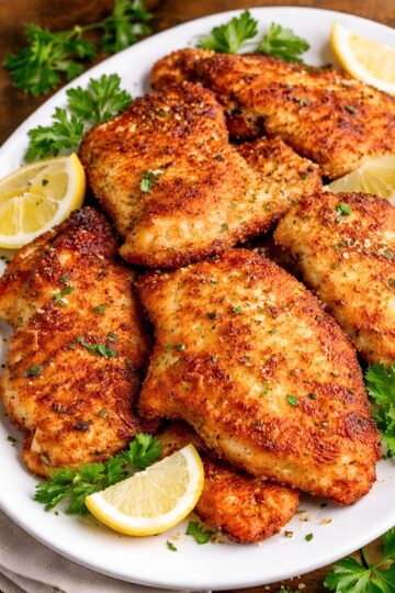 Crispy skillet Parmesan chicken on a white platter with lemon wedges and fresh parsley. Golden brown breaded chicken breasts are arranged close together in a plated food photo with a rustic wood background, showing a crunchy coating and herb garnish.