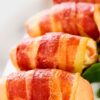 Close-up of bacon wrapped chicken tenders on a white plate, with crispy golden bacon wrapped around juicy chicken and a few green leaves on the side for garnish.