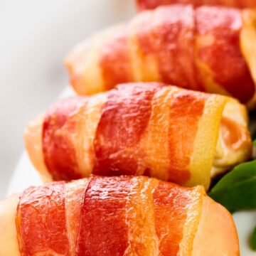 Close-up of bacon wrapped chicken tenders on a white plate, with crispy golden bacon wrapped around juicy chicken and a few green leaves on the side for garnish.