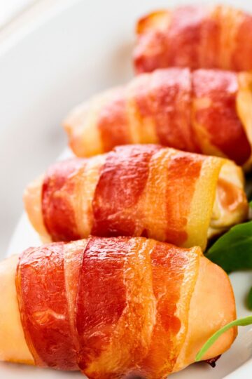 Close-up of bacon wrapped chicken tenders on a white plate, with crispy golden bacon wrapped around juicy chicken and a few green leaves on the side for garnish.