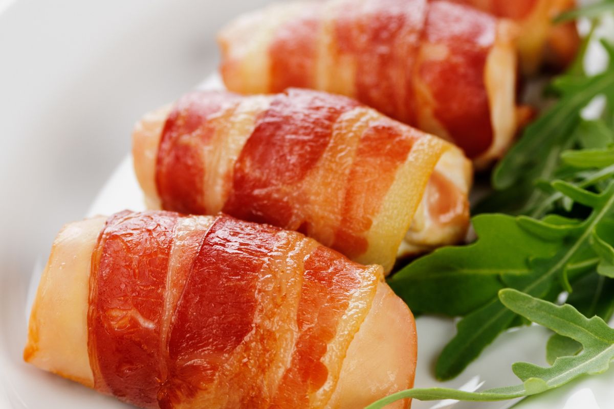 Bacon wrapped chicken tenders on a white plate, showing crisp, golden-brown bacon wrapped around tender chicken, with fresh arugula greens on the side. High protein chicken dinner, easy baked bacon wrapped chicken tenders.