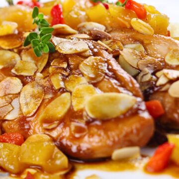 Close-up of baked almond apricot chicken breasts with glossy apricot sauce, sliced toasted almonds, diced apricots, and red pepper on a white plate. Fresh thyme garnish on top, with a soft blurred background.