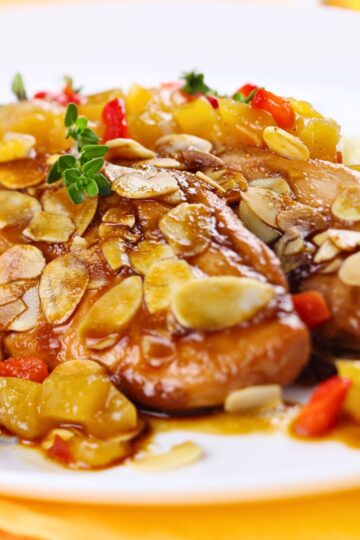 Close-up of baked almond apricot chicken breasts with glossy apricot sauce, sliced toasted almonds, diced apricots, and red pepper on a white plate. Fresh thyme garnish on top, with a soft blurred background.