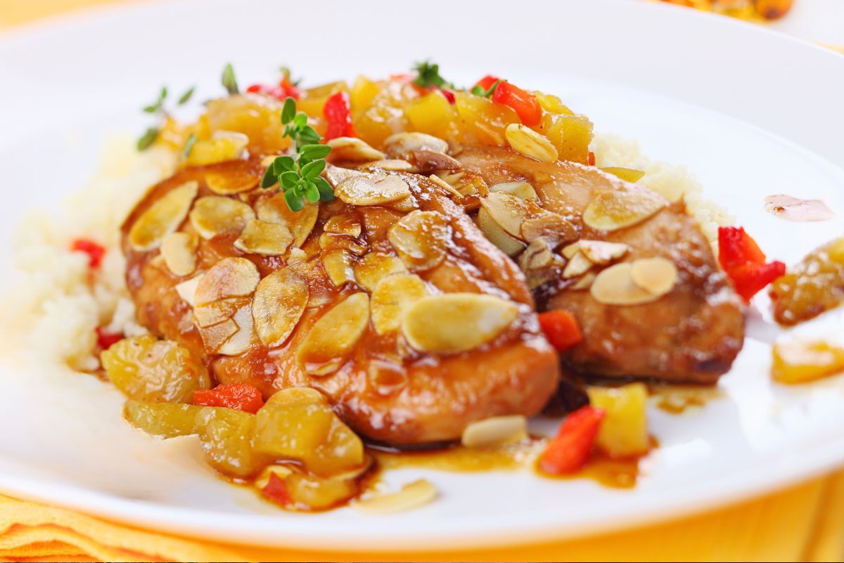 Baked almond apricot chicken breasts on a white plate with rice, glossy apricot sauce, sliced toasted almonds, diced apricots, red pepper, and thyme garnish. A fork rests on the plate, with a yellow napkin underneath and a blurred drink in the background.