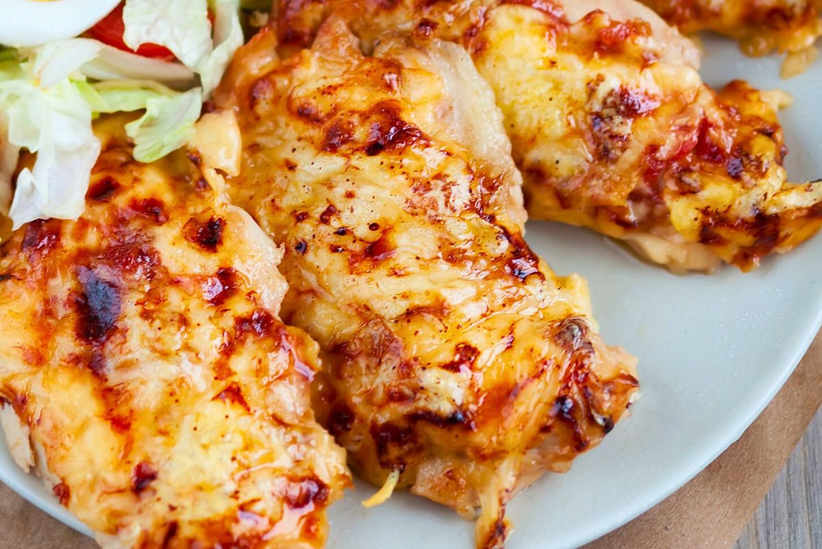 Cheesy baked chicken breasts with melted golden cheese and browned edges on a gray plate. Close-up food photo with juicy baked chicken and a small side salad of lettuce and sliced egg in the corner.