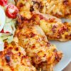 Cheesy baked chicken breasts on a gray plate, topped with melted golden cheese and browned spots. Served with a simple side salad of iceberg lettuce, halved cherry tomatoes, and part of a sliced hard boiled egg. Close-up food photo with a homemade baked chicken dinner presentation.