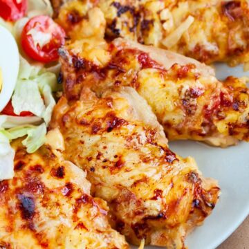 Cheesy baked chicken breasts on a gray plate, topped with melted golden cheese and browned spots. Served with a simple side salad of iceberg lettuce, halved cherry tomatoes, and part of a sliced hard boiled egg. Close-up food photo with a homemade baked chicken dinner presentation.