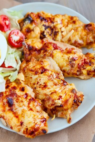 Cheesy baked chicken breasts on a gray plate, topped with melted golden cheese and browned spots. Served with a simple side salad of iceberg lettuce, halved cherry tomatoes, and part of a sliced hard boiled egg. Close-up food photo with a homemade baked chicken dinner presentation.