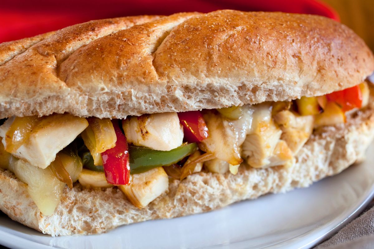 Chicken Philly sub on a whole wheat hoagie roll with grilled chicken, melted cheese, red and green peppers, and sautéed onions on a white plate. Close-up sandwich photo with a red napkin in the background.