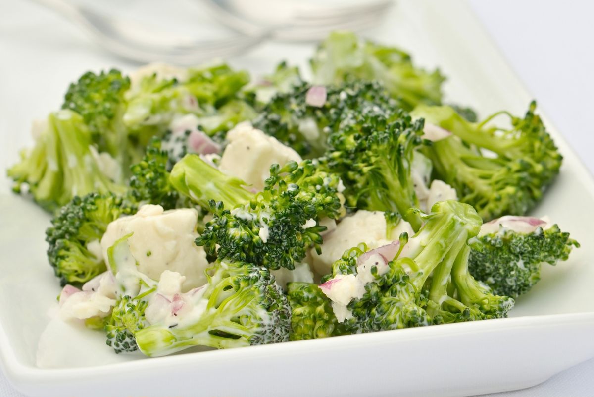 Creamy broccoli and feta salad in a white square bowl with broccoli florets, feta cheese chunks, red onion, and creamy dressing. Fresh healthy salad close-up on a white background.