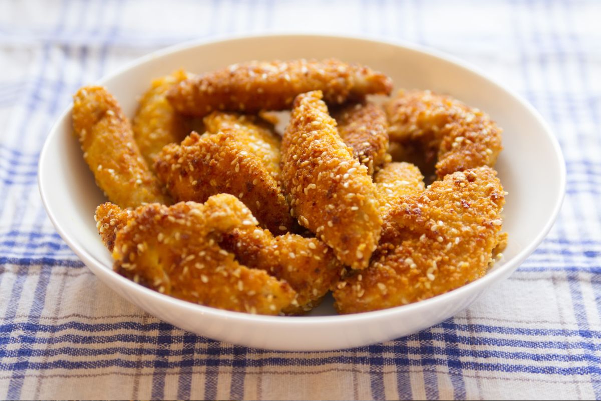 White bowl filled with crispy sesame chicken strips on a blue and white checkered cloth. Golden air fryer chicken tenders have a crunchy breadcrumb coating with sesame seeds.