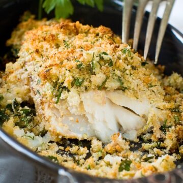 Close-up of flaky baked cod in a small cast-iron skillet, topped with a golden herb-and-garlic parmesan breadcrumb crust. A fork is stuck in the fish, with a parsley sprig in the background and toasted crumbs scattered around for a crispy baked cod dinner photo.