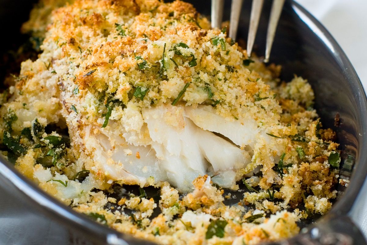 Close-up of flaky baked cod in a small cast-iron skillet, covered with crispy herb, garlic, and parmesan breadcrumb topping. Golden toasted crumbs and chopped green herbs are scattered around, with the tips of a fork visible at the top for a savory baked cod dinner photo.