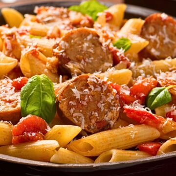 Rigatoni pasta with sliced Italian sausage, cherry tomatoes, fresh basil, and grated Parmesan in a rich tomato sauce, served in a dark bowl. Warm, cozy weeknight dinner pasta with a close-up food photography style.
