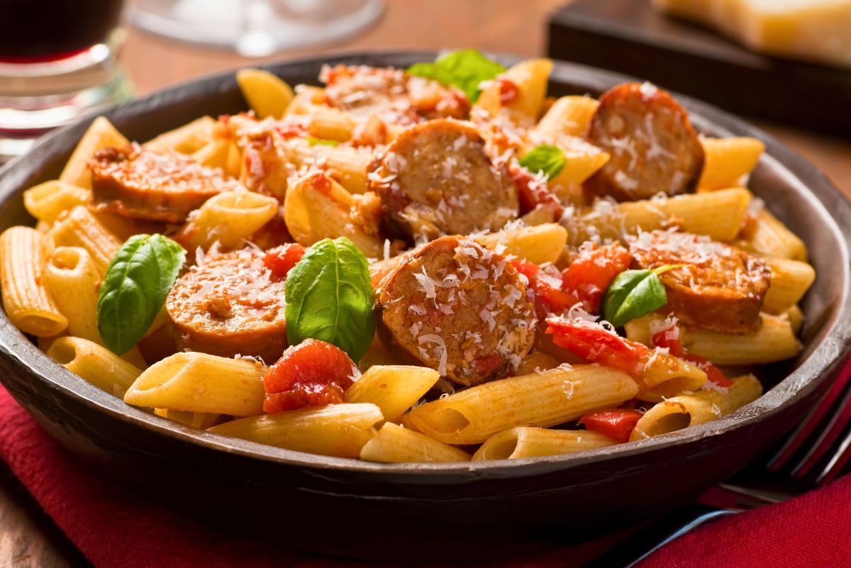 Bowl of rigatoni with sliced Italian sausage, cherry tomatoes, fresh basil, and grated Parmesan, served in light tomato sauce. Cozy pasta dinner on a red napkin with a drink in the background. High protein Italian sausage pasta and easy weeknight dinner food photography.