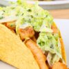 Close-up of a crispy fish taco in a crunchy corn shell, filled with seasoned fish pieces and creamy lime slaw on a white plate. Blurred background shows a bowl of red tomatoes