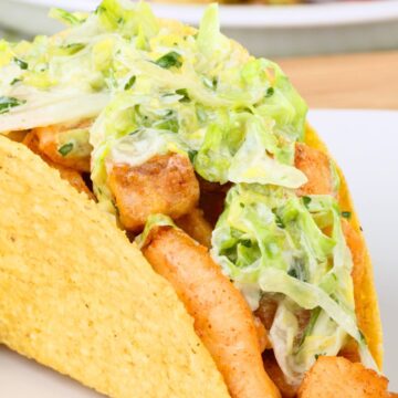 Close-up of a crispy fish taco in a crunchy corn shell, filled with seasoned fish pieces and creamy lime slaw on a white plate. Blurred background shows a bowl of red tomatoes