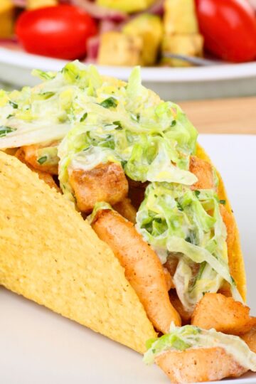 Close-up of a crispy fish taco in a crunchy corn shell, filled with seasoned fish pieces and creamy lime slaw on a white plate. Blurred background shows a bowl of red tomatoes