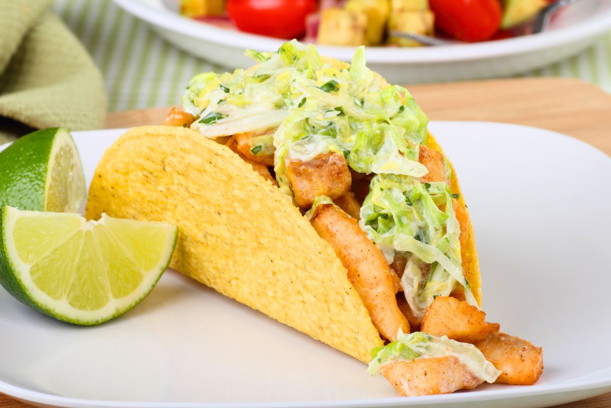 Crispy fish tacos with creamy lime slaw on a white plate, served with fresh lime wedges. Crunchy taco shells are filled with seasoned fish pieces and shredded slaw. Blurred background shows a bowl with tomatoes