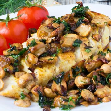 Garlic herb chicken breast topped with sautéed mushrooms, caramelized onions, and chopped green herbs on a white plate, with three vine tomatoes on the side. Rustic wood table background with fresh dill and a fork in the corner.